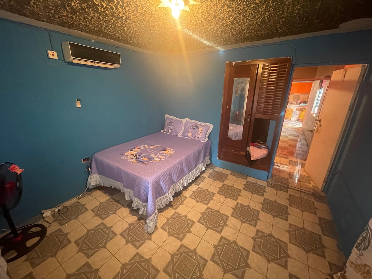 A room features a spacious bed covered with a purple bedspread and decorative pillows. Blue walls create a calming environment, and a window allows natural light to illuminate the space. An air conditioning unit and a small fan are visible, enhancing comfort.