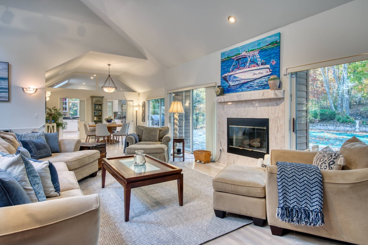 An open living space features a comfortable arrangement of neutral-toned sofas, a wooden coffee table, and a light area rug. Natural light fills the room through large glass doors, with a fireplace framed in stone and artwork displayed above, enhancing the relaxing ambiance.