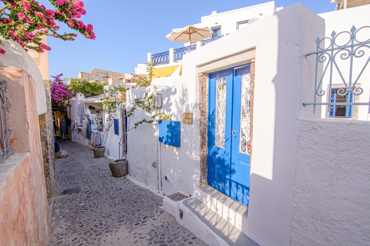 A charming alleyway features a whitewashed structure with bold blue doors, leading into the inviting home. Vibrant bougainvillea blooms add splashes of color along the stone pathway, while a terrace is visible above, complemented by an umbrella providing shade.