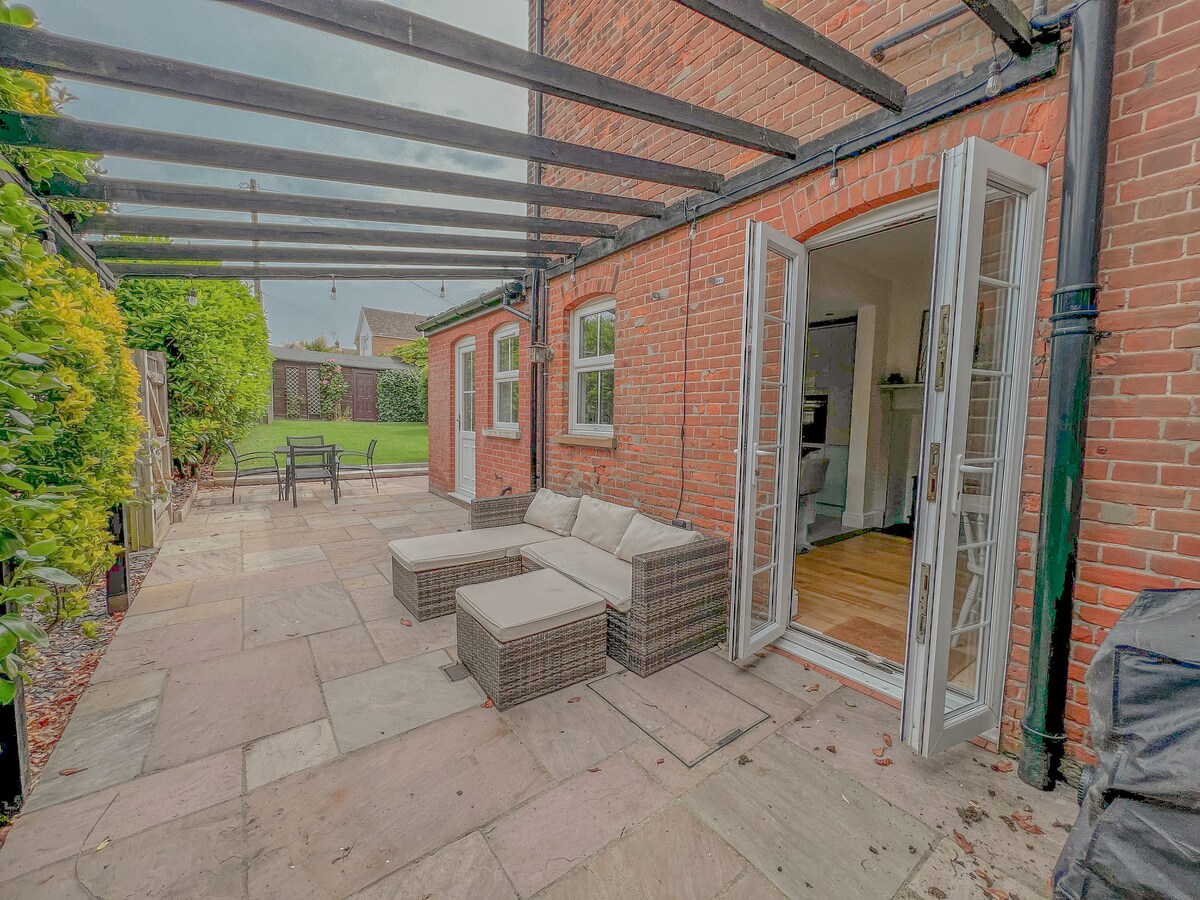 An outdoor patio area is visible, featuring a seating arrangement with soft cushions. French doors lead into the interior space, with a view of the garden beyond. The stone flooring complements the brick exterior, creating a harmonious transition between indoor and outdoor living.