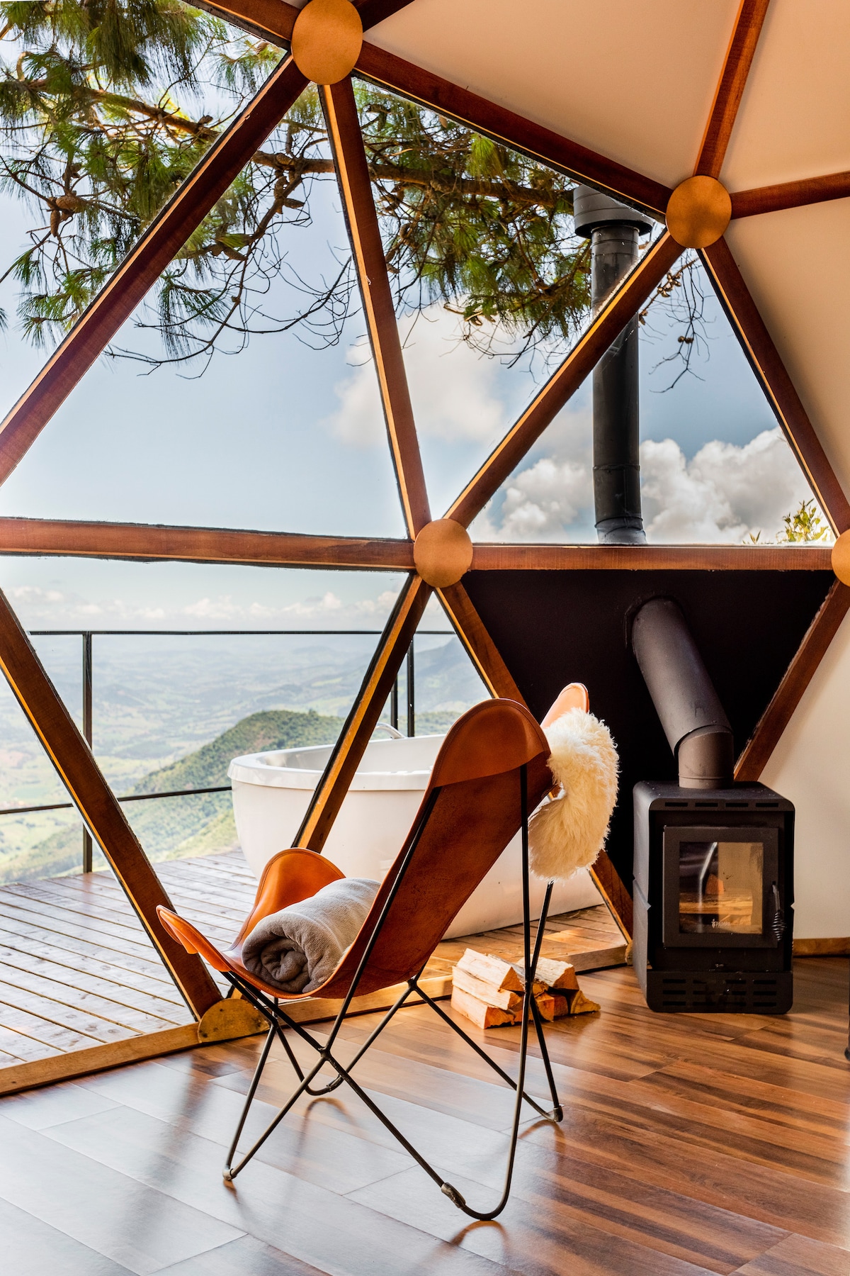 A cozy seating area features a butterfly chair and a small blanket, complemented by a wood stove. Large windows framed by geometric designs provide expansive views of the surrounding nature and mountains. A white bathtub is positioned on the wooden deck, inviting relaxation.