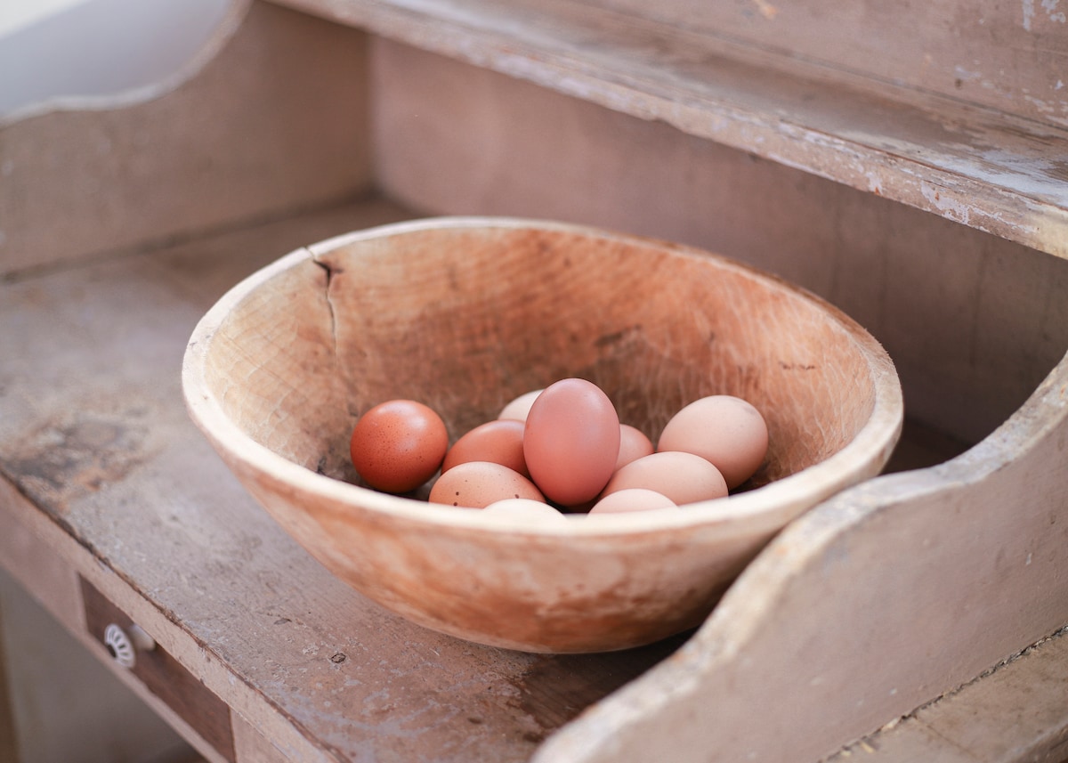 A wooden bowl rests on a weathered shelf, filled with several eggs in varying shades of brown. The natural texture of the bowl complements the rustic charm of the setting.