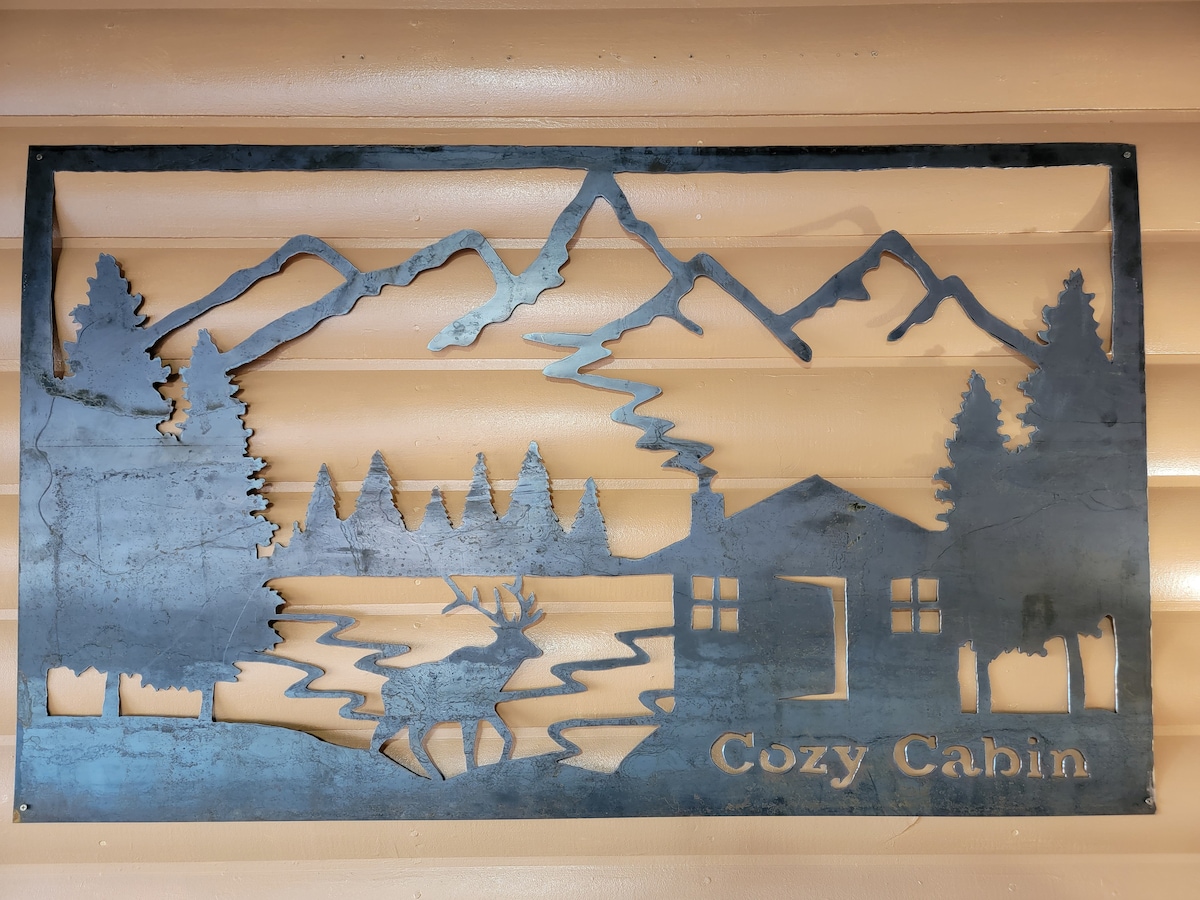 A metal art piece depicting a mountain landscape, featuring tall pine trees, a cabin, and a deer beside a river. The words 'Cozy Cabin' are inscribed at the bottom, all set against a wooden wall.