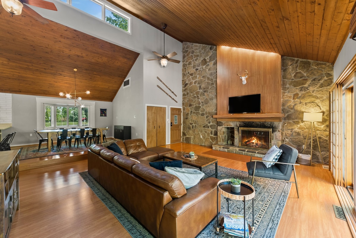 A spacious living area features a vaulted wood ceiling above a large leather sectional sofa. A stone fireplace is prominently displayed, complemented by a TV mounted above. Natural light floods the space, with large doors leading to the outdoor deck visible in the background.