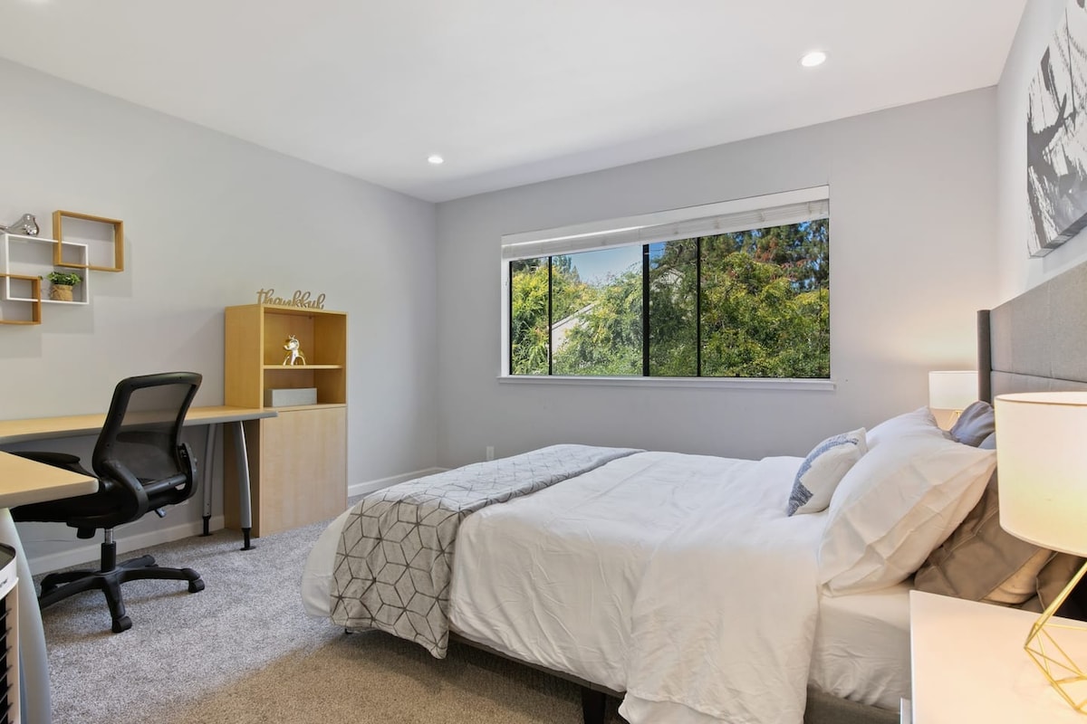 A well-lit bedroom features a spacious bed dressed in white linens, complemented by a soft gray throw. A large window presents a view of greenery. A desk with a chair and a wooden shelf are positioned against the wall, providing a cozy workspace.