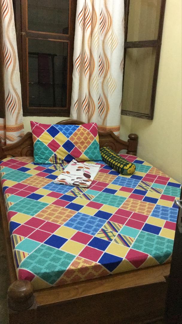 A cozy sleeping area features a patterned bedspread in vibrant colors, complemented by a square pillow. A rolled pillow adds a touch of comfort. Flowing curtains with a wavy design frame a window, allowing natural light to brighten the space.