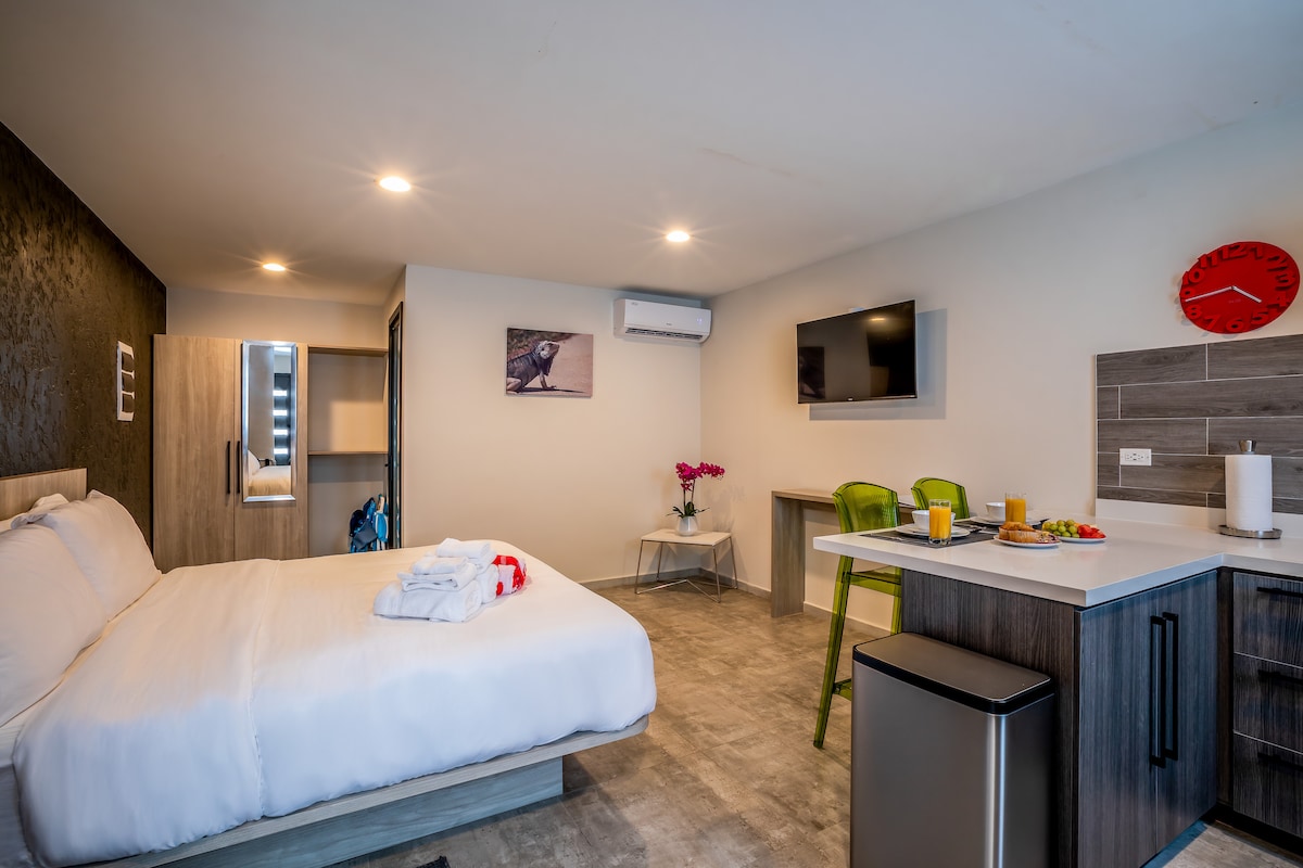 A bright studio apartment features a spacious bed with fresh linens and decorative towels folded neatly on top. A small dining table and vibrant green chairs complement the sleek kitchen area, equipped with modern appliances. A wall-mounted TV is visible, alongside contemporary lighting fixtures.