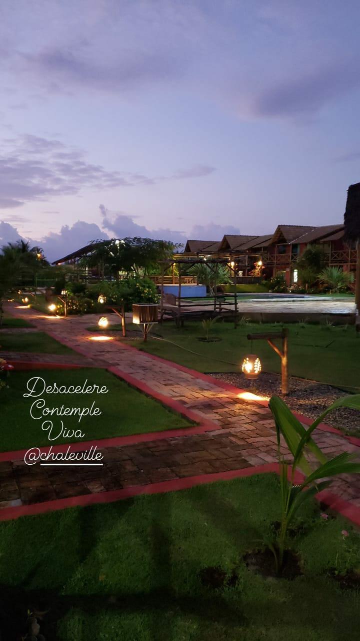 A scenic pathway is lined with soft lighting and surrounded by lush greenery. The pathway leads to various amenities, including a pool visible in the background. Traditional structures with thatched roofs are situated along the path, creating a serene atmosphere as twilight sets in.