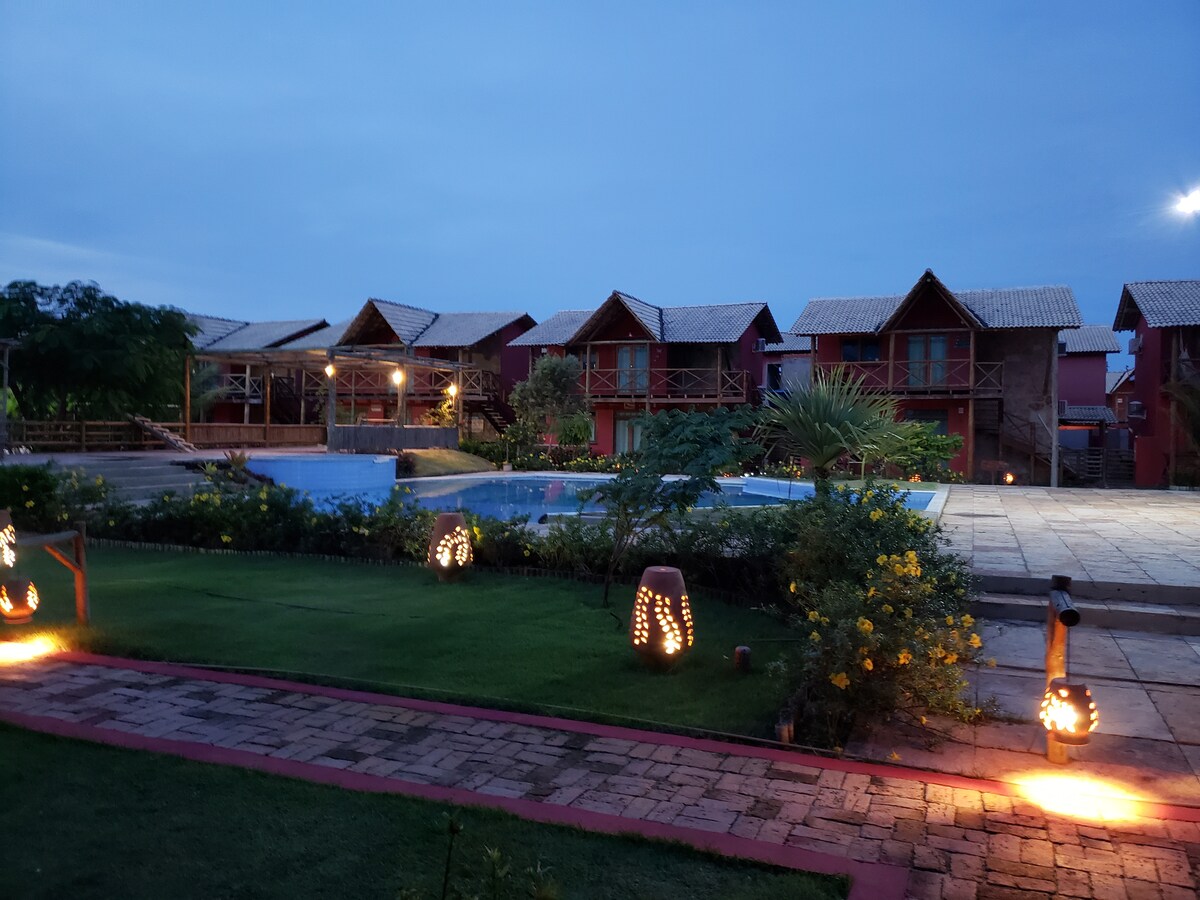 The image captures a serene evening scene, featuring a well-lit pool area surrounded by multiple wooden chalets. Soft lighting highlights the landscaped garden, while stone pathways lead through the lush greenery, creating a tranquil setting for relaxation.