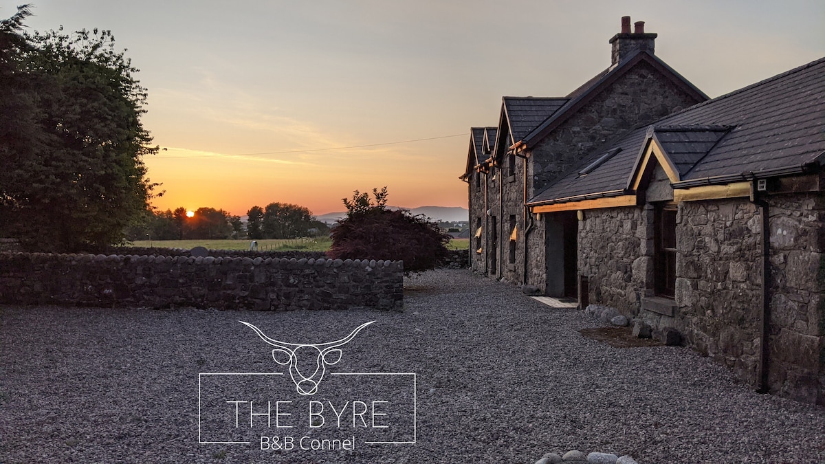 The exterior of The Byre B&B is captured during sunset, highlighting the stone building and surrounding gravel area. The warm light from the setting sun casts a soft glow, while the tree-lined background adds depth to the serene rural landscape.