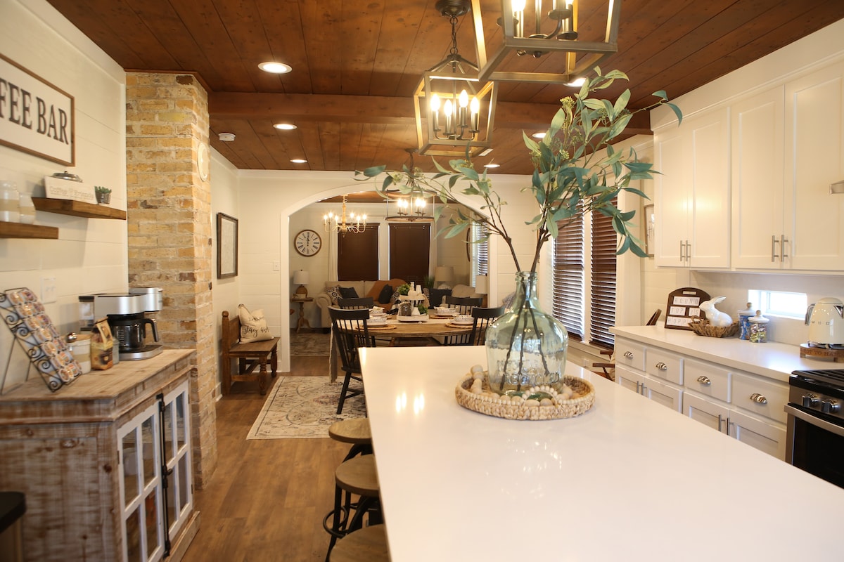 A modern kitchen features new stainless steel appliances and a spacious island with seating. A coffee bar is positioned nearby, surrounded by white cabinetry and a rustic wood accent wall. Natural light enters through windows, providing a warm and inviting ambiance.
