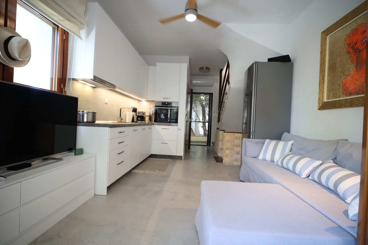 A modern kitchen area features sleek white cabinetry and stainless steel appliances. A spacious living area includes a light-colored sofa and a large flat-screen TV. Natural light enters through large windows, enhancing the contemporary design.