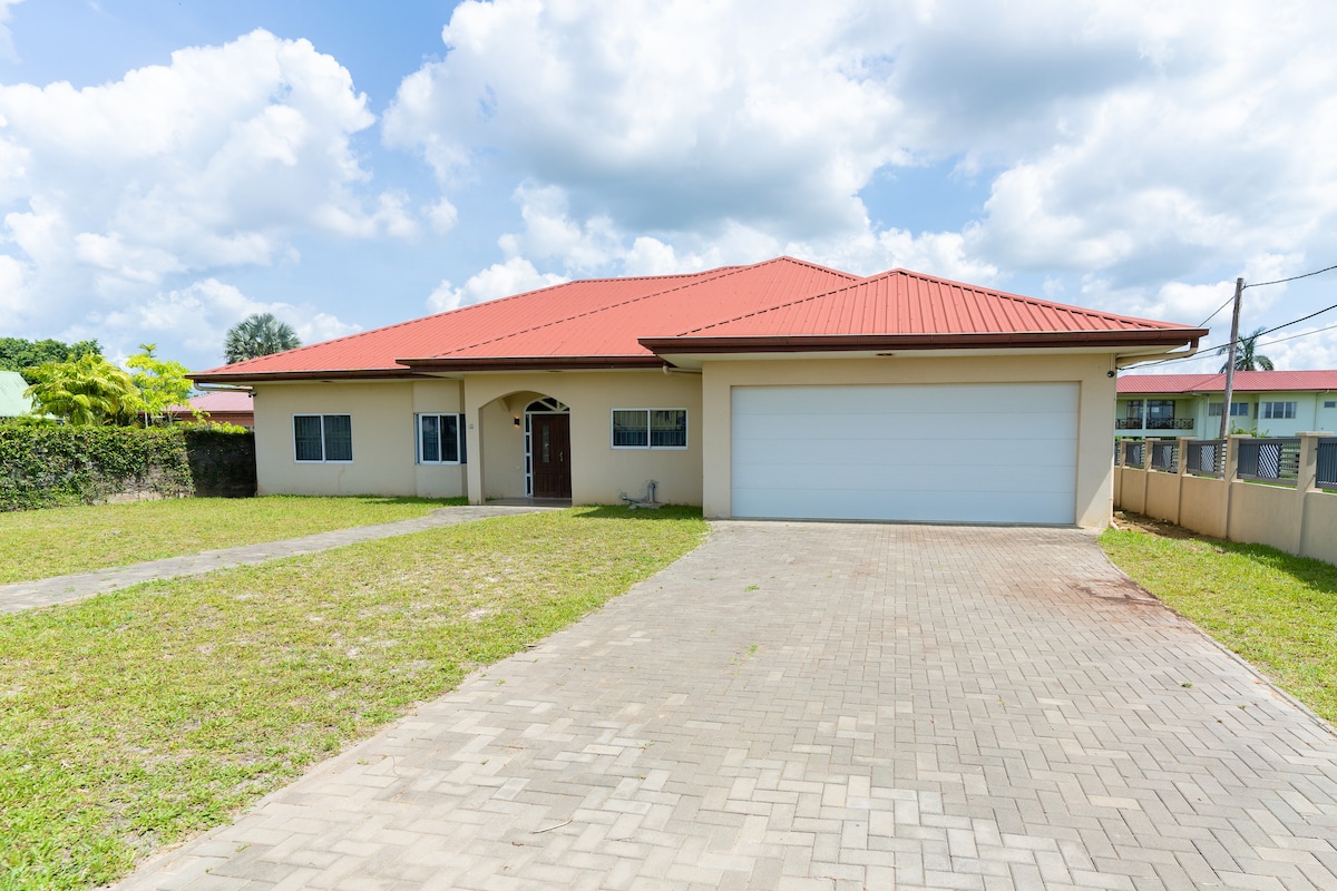 The villa is shown featuring a spacious front yard with well-maintained grass. A large driveway leads to a two-car garage with a flat roof. The exterior is complemented by a red tiled roof and multiple windows, surrounded by greenery and under a partly cloudy sky.