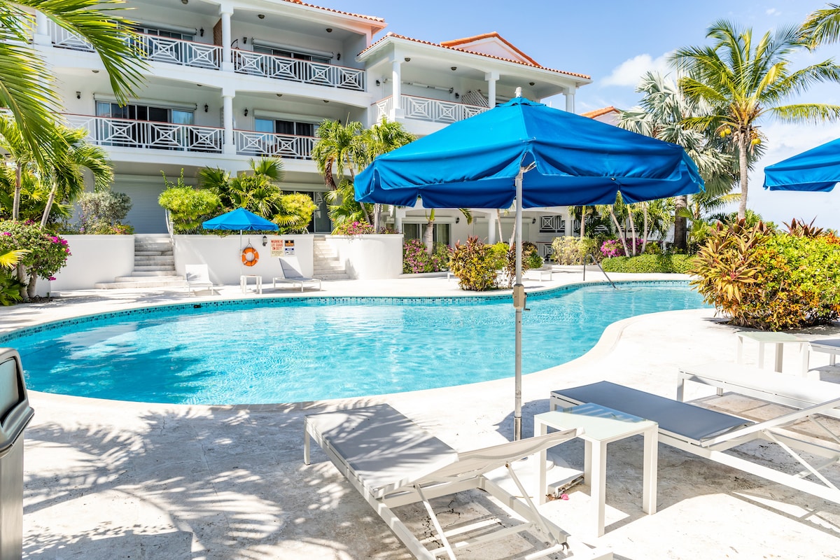 A serene pool area is surrounded by lush tropical landscaping and palm trees. Blue umbrellas provide shade over lounge chairs positioned near the clear water. The resort architecture features multiple levels, with balconies framed by vibrant flowers and greenery.