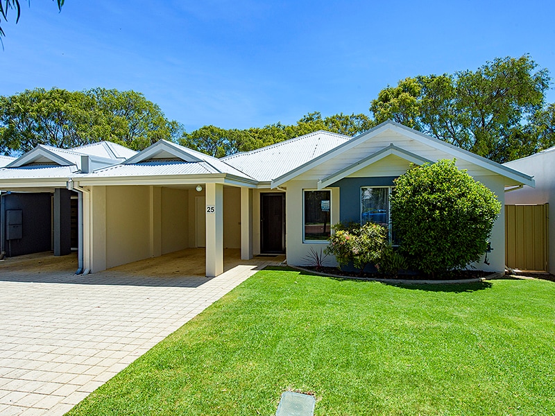 A single-story cottage is displayed, featuring a welcoming façade with a covered entrance. Lush green grass surrounds the property, and well-maintained shrubs are positioned near the windows. The driveway is paved, leading to a clear view of the home's inviting exterior against a blue sky.