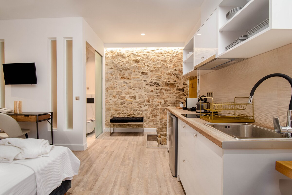 A modern studio space features a combination of natural stone and sleek cabinetry. An open kitchenette with a sink and countertop is positioned beside a dining area. A comfortable sleeping area is visible, with light wood flooring enhancing the overall brightness of the room.