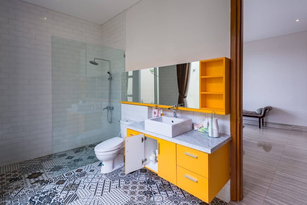 A modern bathroom is featured with a spacious shower area enclosed by clear glass. A sink with a marble countertop sits beneath a large mirror. Colorful cabinetry in orange provides storage, and patterned tiles add visual interest to the floor.
