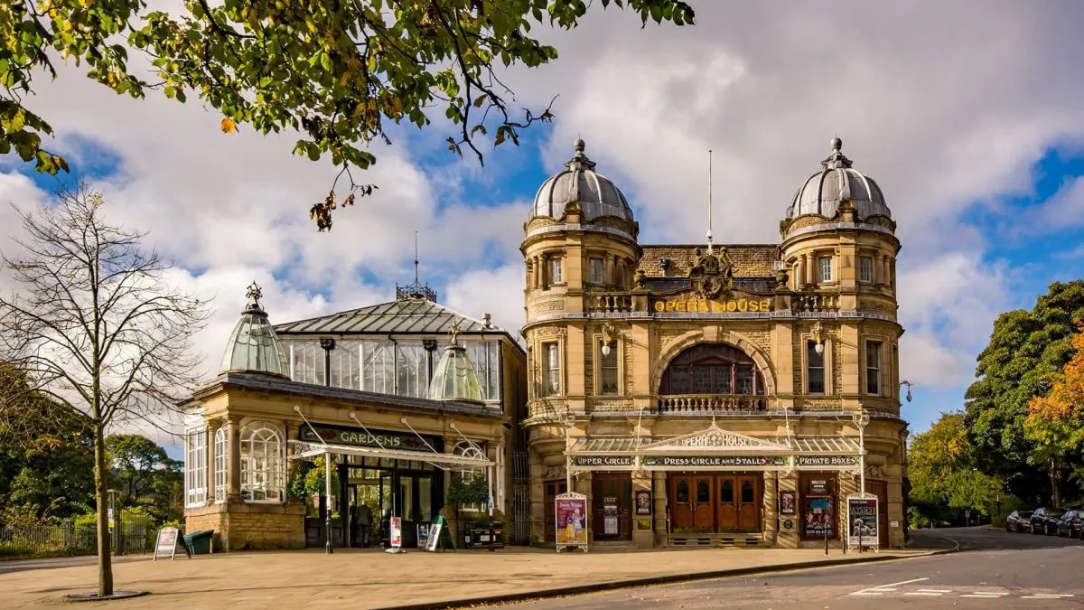 The image captures a historic theater building known for its ornate structure and grand facade. The entrance showcases intricate architectural details, with large windows and a domed roof. Surrounding trees provide greenery, and a clear sky complements the vibrant atmosphere.