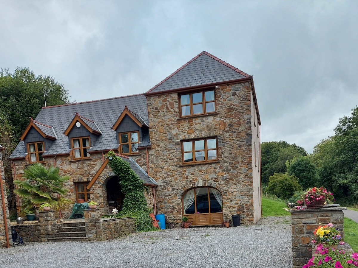 A spacious stone exterior showcases a two-story home with multiple windows, allowing natural light to illuminate the warm, earthy tones of the structure. Lush greenery and colorful planters line the pathway, leading to a gravel driveway that provides ample parking space.