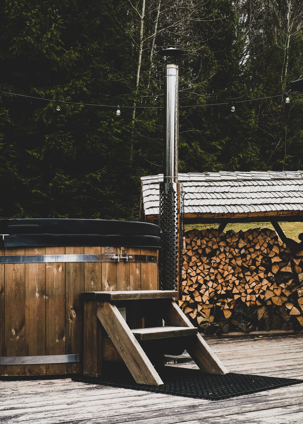 A wooden hot tub is positioned on a deck with a sturdy set of steps leading up to it. A chimney is attached to the hot tub, and a wooden shelter with stacked logs is visible in the background. Lush greenery surrounds the area.