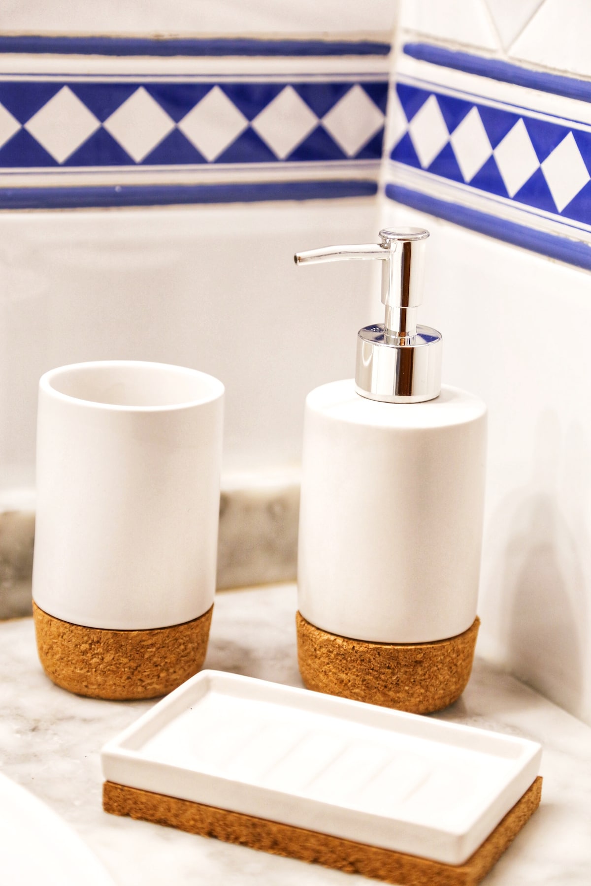 A minimalist bathroom arrangement is presented, featuring two white containers with cork bases. One container serves as a soap dispenser with a sleek metallic pump, while the other is a tumbler. A rectangular soap dish sits nearby, also with a cork base.