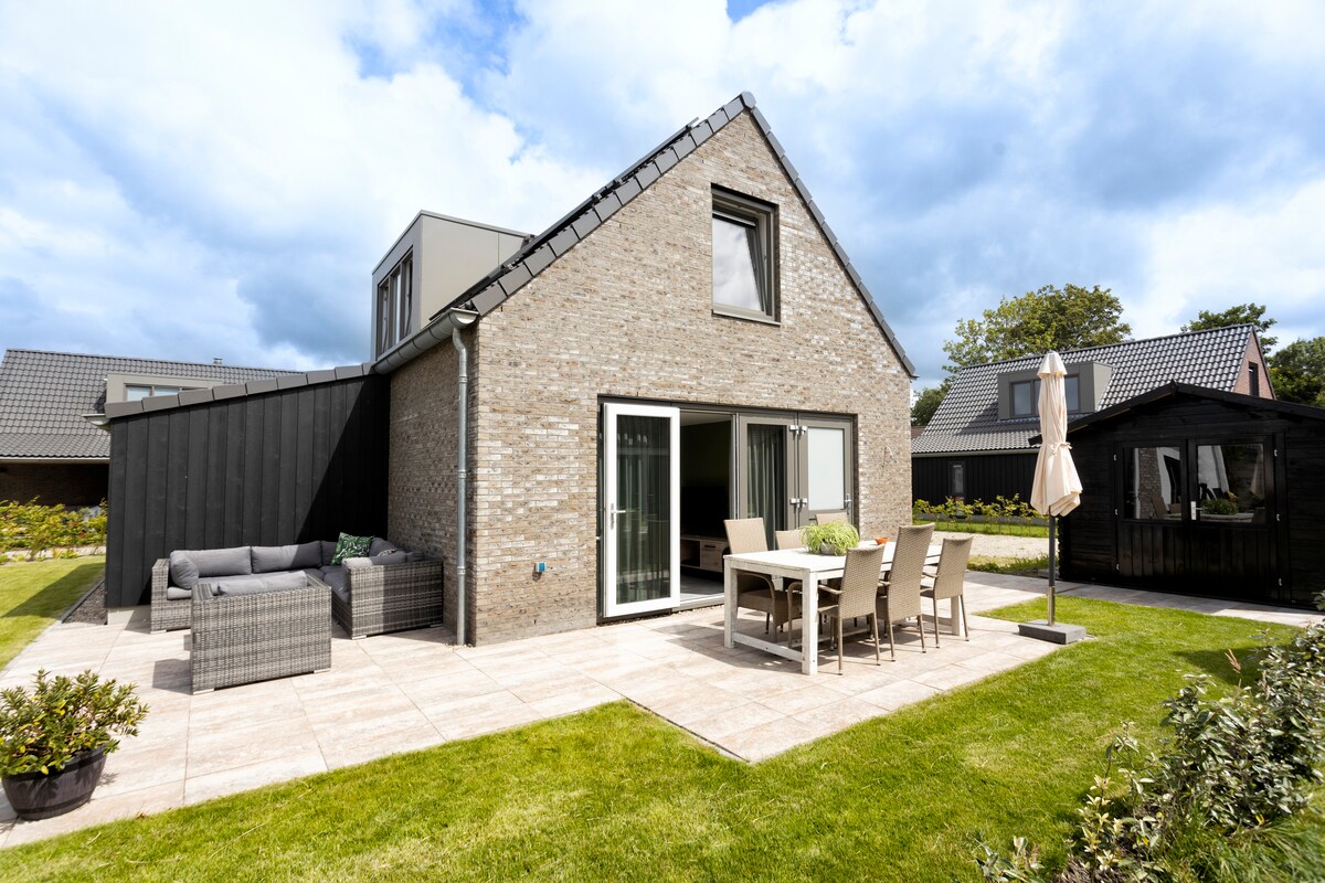 The exterior of the vacation villa is showcased, featuring a light-colored brick facade. A spacious terrace is equipped with a dining table and chairs, alongside a lounge area. Lush green lawn surrounds the property, and a cozy garden shed is visible in the background.