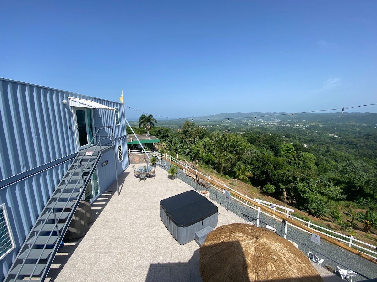 An open terrace showcases a relaxing space with a hot tub and a thatched umbrella for shade. Stairs lead to an upper level, while panoramic views of the surrounding landscape extend to the horizon under a clear blue sky.