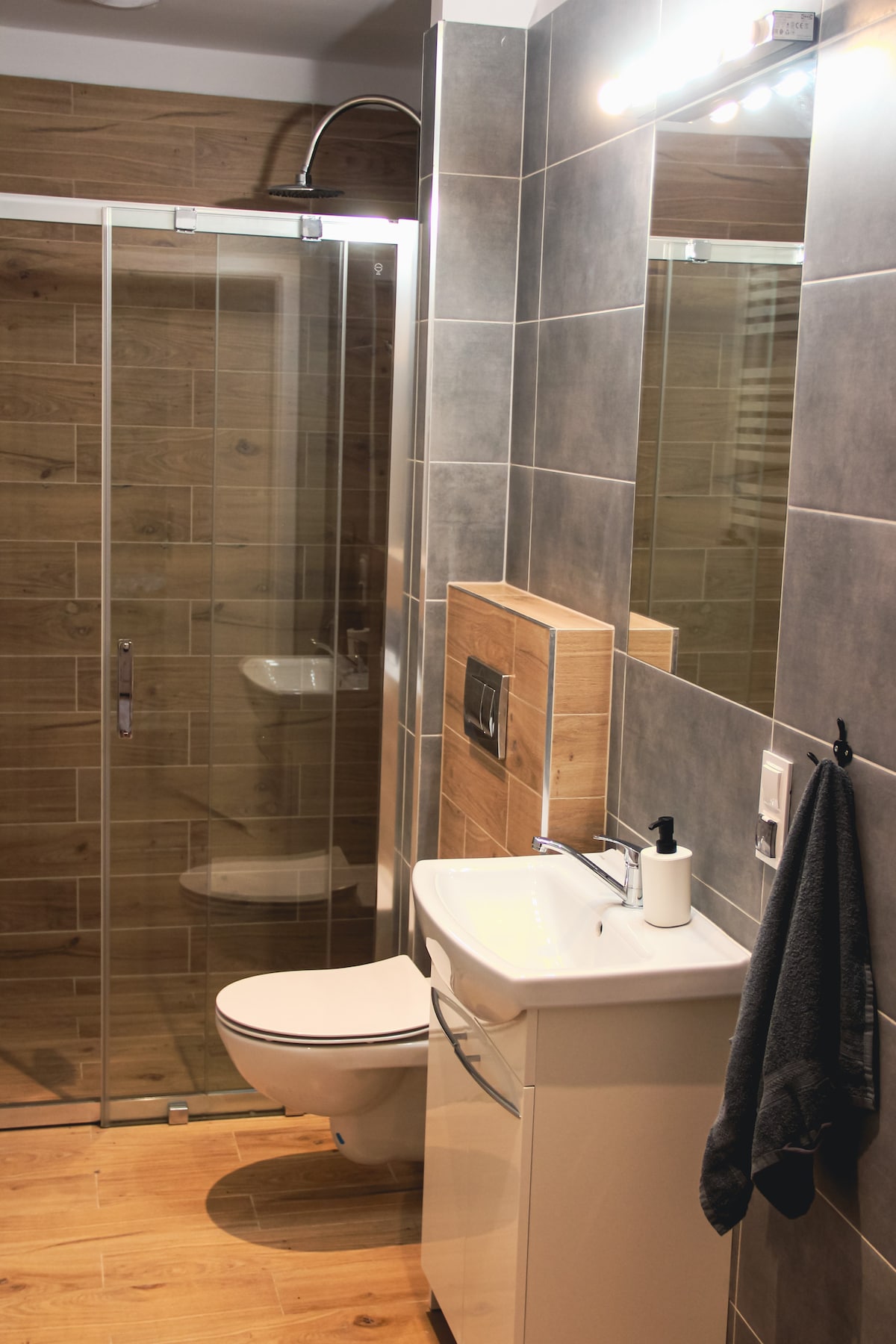A modern bathroom is presented, featuring a walk-in shower enclosed by glass panels. A sleek sink and vanity are positioned next to the shower, with wooden accents on the walls. A full-length mirror reflects the bright lighting from above, and a towel is neatly hung nearby.