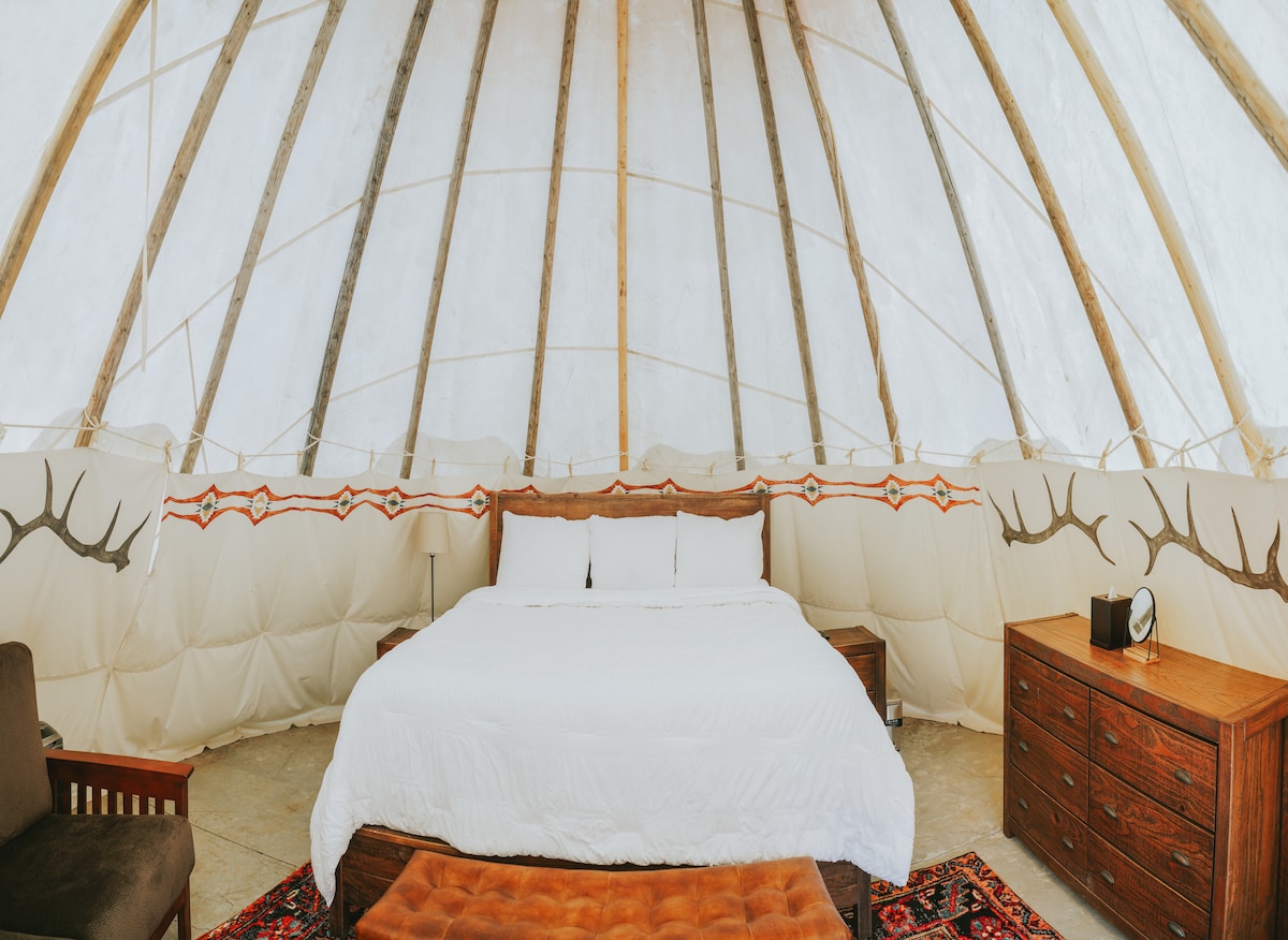 The interior of the Tipi features a spacious bed with white linens and decorative pillows. Natural wood elements are present, along with a patterned rug. Nearby, bedside tables and a clock are visible, contributing to the cozy atmosphere of the space.