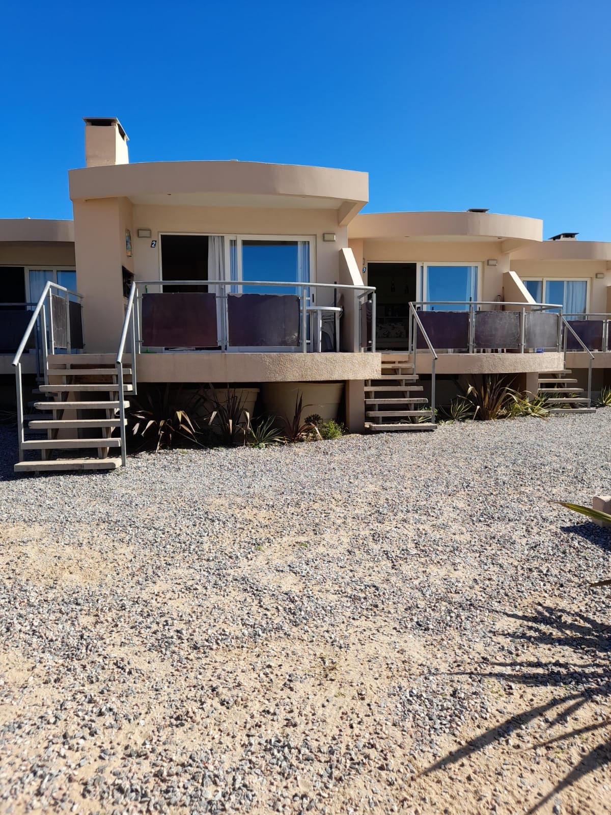 Two individual apartment units are showcased in this image, featuring modern architecture with rounded balconies. Each unit has large glass doors leading to the outdoors. A gravel pathway and neatly arranged plants enhance the exterior landscape under a clear blue sky.