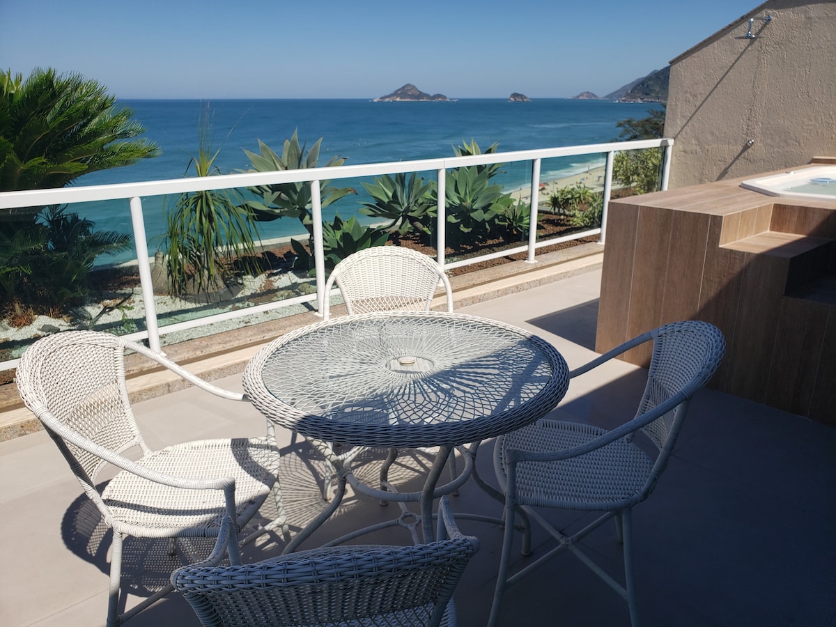 A terrace is presented with a round table surrounded by four white wicker chairs. The backdrop features a sweeping ocean view, highlighting distant islands against a clear blue sky. Lush greenery frames the scene, enhancing the serene coastal ambiance.