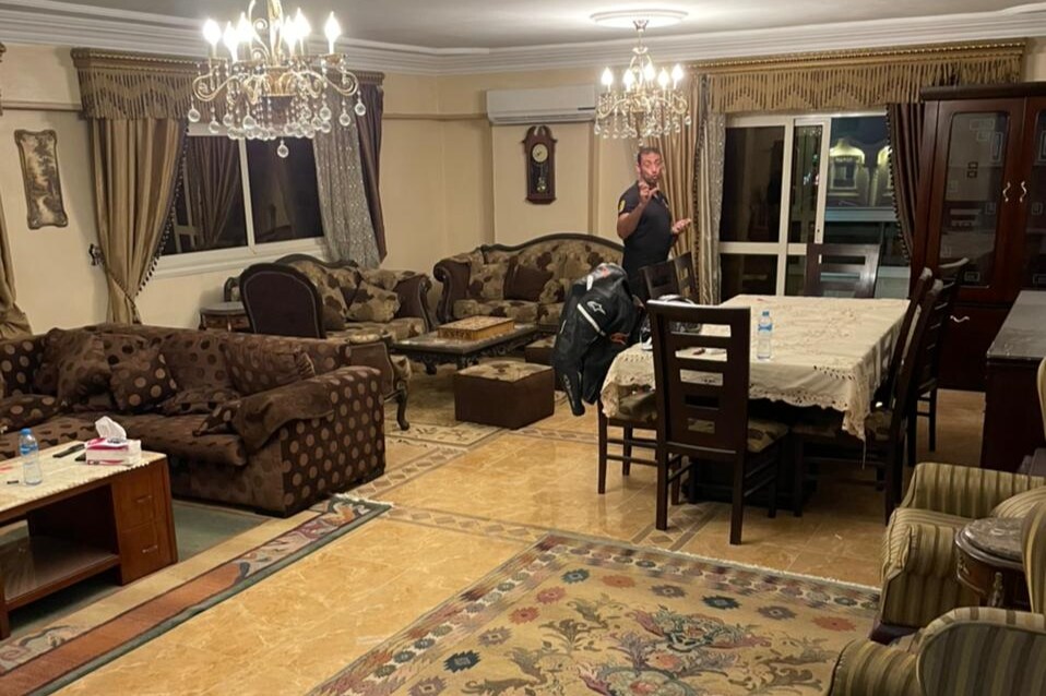 The spacious living area features a mix of elegant furniture, including patterned sofas and a formal dining table. Two chandeliers provide illumination, while large windows allow natural light to enter. A decorative rug adds warmth to the floor, enhancing the inviting ambiance.