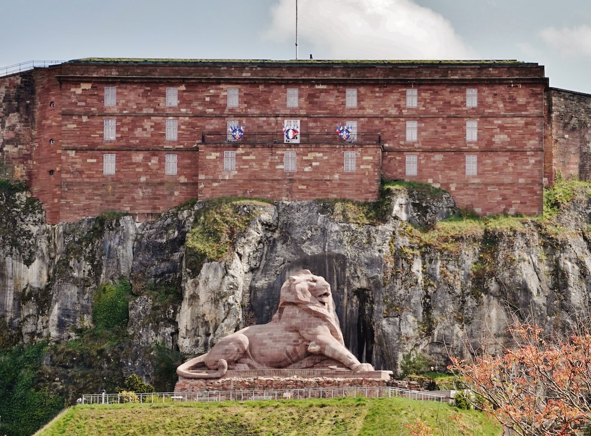 A large stone lion sculpture is positioned at the base of rocky hills, depicting a majestic posture. Above, a historic fortress is seen, constructed with reddish stone and featuring multiple windows. The scene is set against a backdrop of green foliage and a partly cloudy sky.