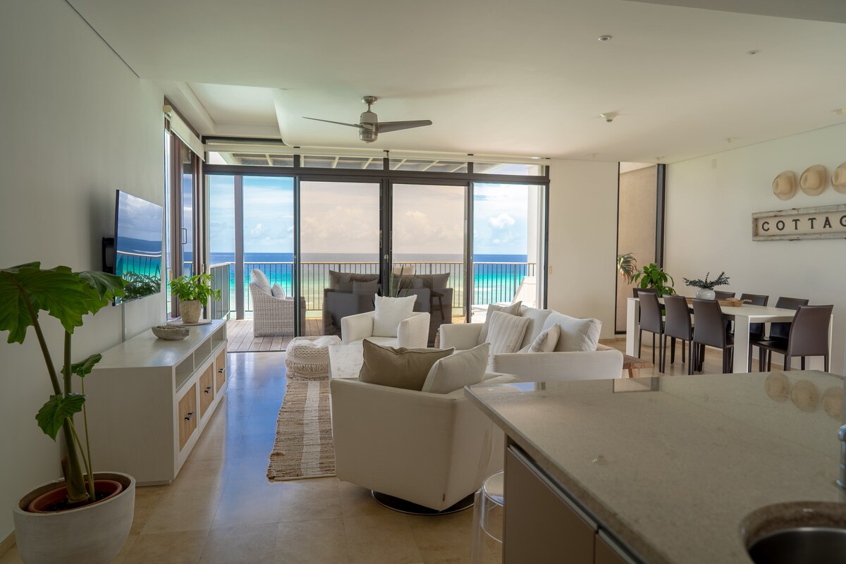 A spacious living area features large glass doors offering views of the lagoon. Comfortable seating arrangements are present, complemented by a ceiling fan and a dining area with a table and chairs. Natural light illuminates the neutral décor, enhancing the serene atmosphere.