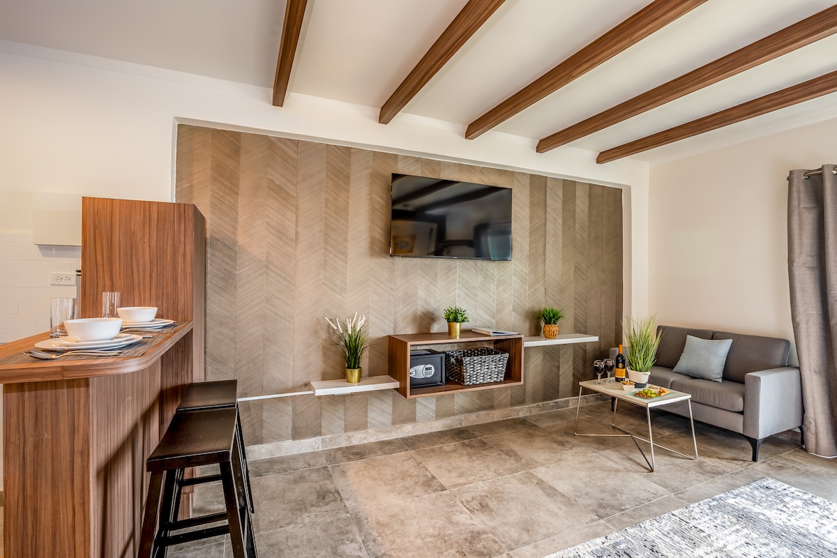 A cozy studio space features an open layout with a modern design. Warm wooden beams span the ceiling. A wall-mounted flat-screen TV is positioned above a sleek media console, complemented by decorative plants. A small dining area is visible with black stools arranged around a light wooden counter.