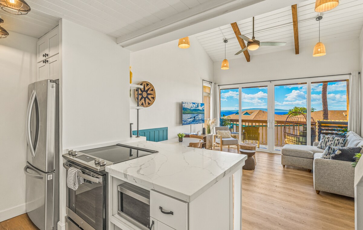 A modern kitchen is visible on the left, featuring stainless steel appliances and a white marble countertop. On the right, a comfortable living area is presented with a light grey sofa and large glass doors offering a view of the ocean and surrounding landscape.