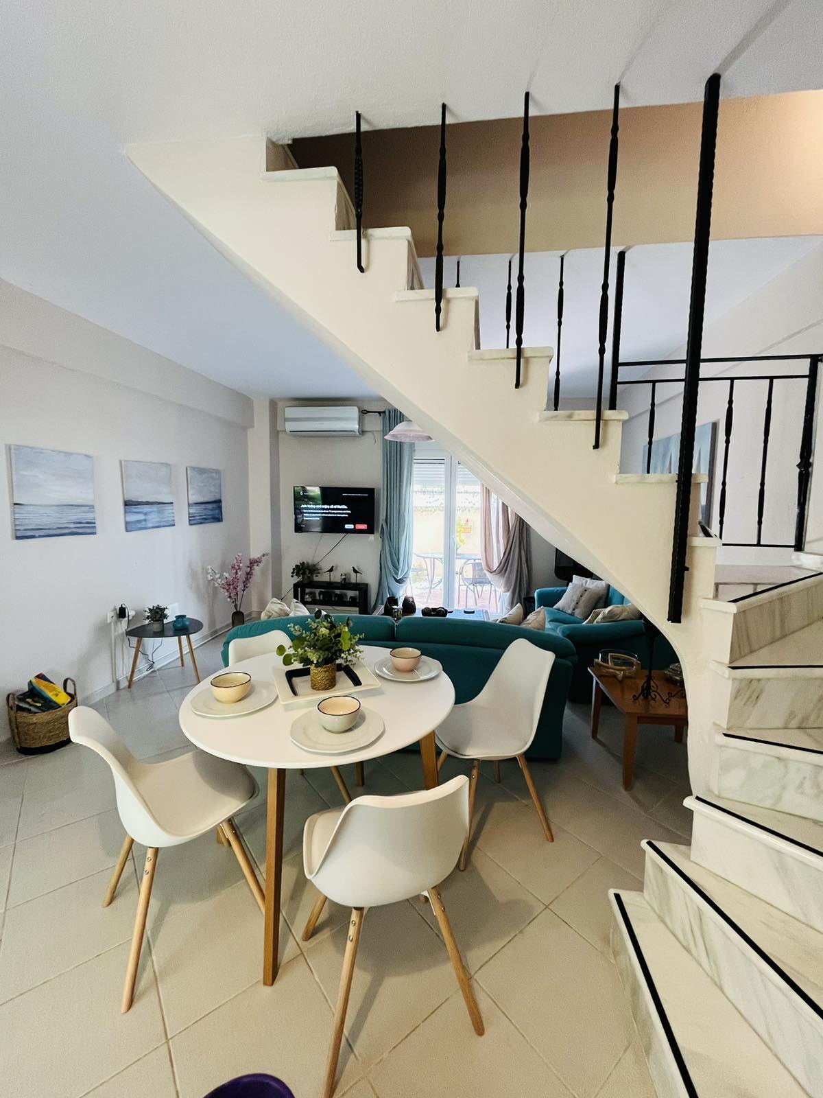 A bright dining area is situated beside a staircase, featuring a round table with four white chairs. Natural light enters through a nearby window, illuminating the room which includes a teal sofa and a wall-mounted television. Decorative elements and plants add a home-like touch.