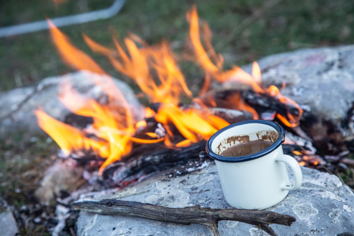 A campfire is depicted with bright flames consuming wood, set against a backdrop of natural surroundings. A metal mug rests on a rock nearby, showcasing the comforts of outdoor living. The scene reflects a tranquil moment in nature.