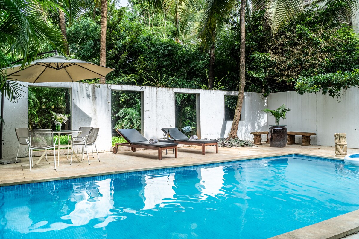 A private swimming pool is surrounded by lush greenery, featuring two lounge chairs and an umbrella for shade. A set of outdoor dining furniture complements the tranquil setting, where tall trees provide a natural backdrop against the white walls.