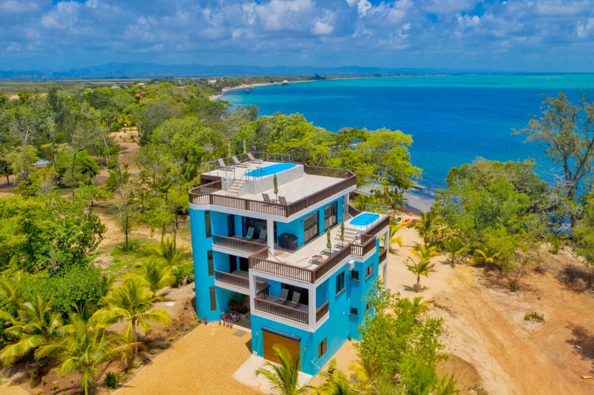 A three-story building in a vibrant blue hue is surrounded by lush greenery and palm trees. The rooftop area features two pools, offering panoramic views of the Caribbean coastline and clear blue waters, creating a tranquil environment.