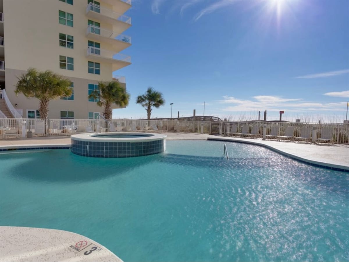 The pool area features a large, inviting turquoise swimming pool with a connected hot tub. Surrounding palm trees provide shade, while the bright blue sky enhances the tranquil atmosphere. The pool deck is equipped with safety railings and lounge chairs for relaxation.