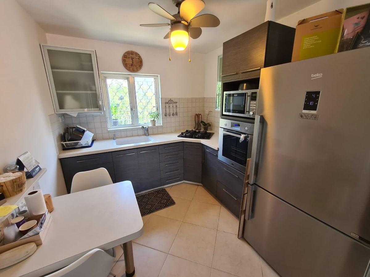 A modern kitchen is equipped with a sleek refrigerator and built-in oven. Light-colored cabinets and a compact table enhance functionality. Large windows allow natural light, while a ceiling fan adds air circulation. Various kitchen utensils and appliances are neatly arranged for convenience.