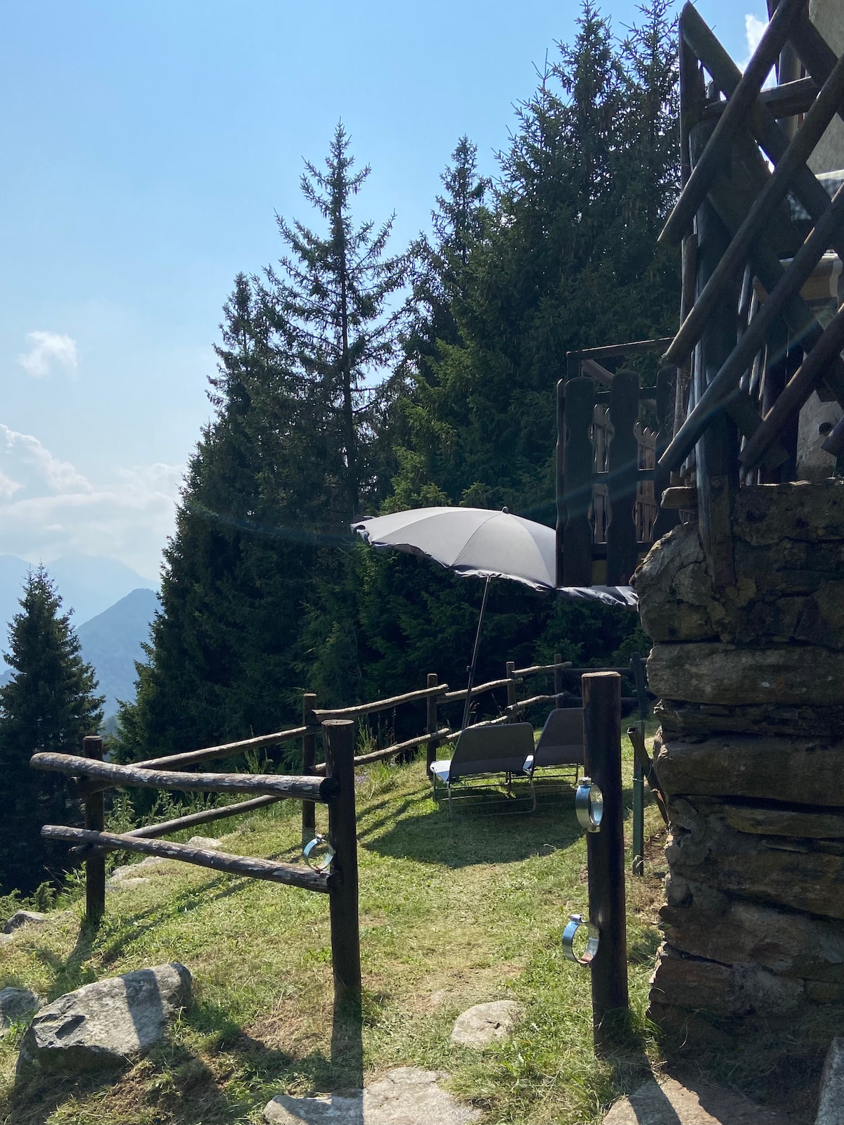 A serene outdoor area features a shaded lounge space under a large umbrella. The grassy area is bordered by rustic wooden fencing, with tall trees providing a natural backdrop. The distant mountains can be seen, enhancing the tranquil atmosphere.