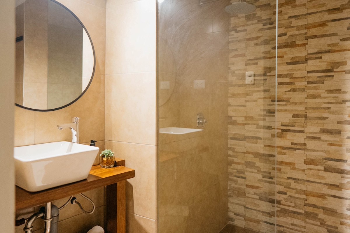 A modern bathroom is presented with warm-toned tiles and a glass shower enclosure. A round mirror hangs above a wooden vanity featuring a square sink. A small potted plant adds a touch of greenery to the serene space.