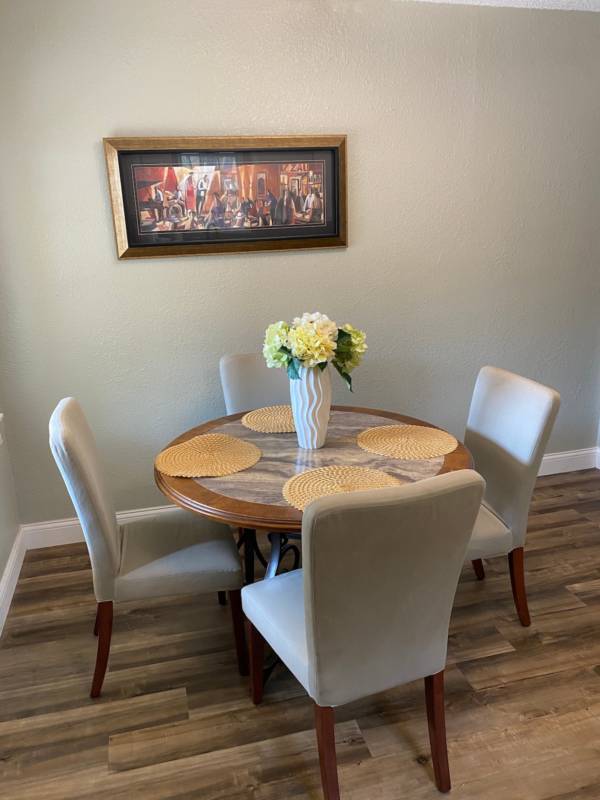 A round dining table is set for four in a well-lit space. Neutral-toned chairs provide comfortable seating. A vase filled with flowers adds a touch of color, and artistic wall decor enhances the dining area.