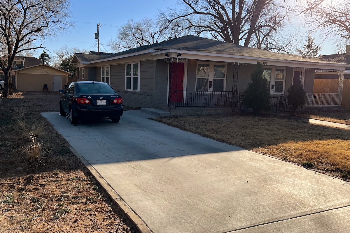 A single-story home with a light-colored façade is set on a spacious lot. A driveway leads to a parked car, while a red front door adds a pop of color. The surrounding area features brown grass and a few trees, contributing to a tranquil outdoor setting.