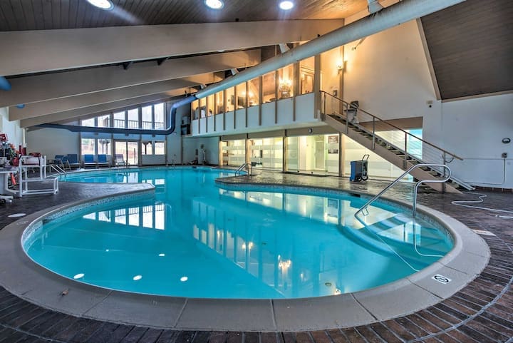 An indoor swimming pool is featured, with clear blue water reflecting the overhead lights. A sloped entryway is visible on one side, and large windows provide natural light. Surrounding lounge chairs are positioned along the pool's edge.