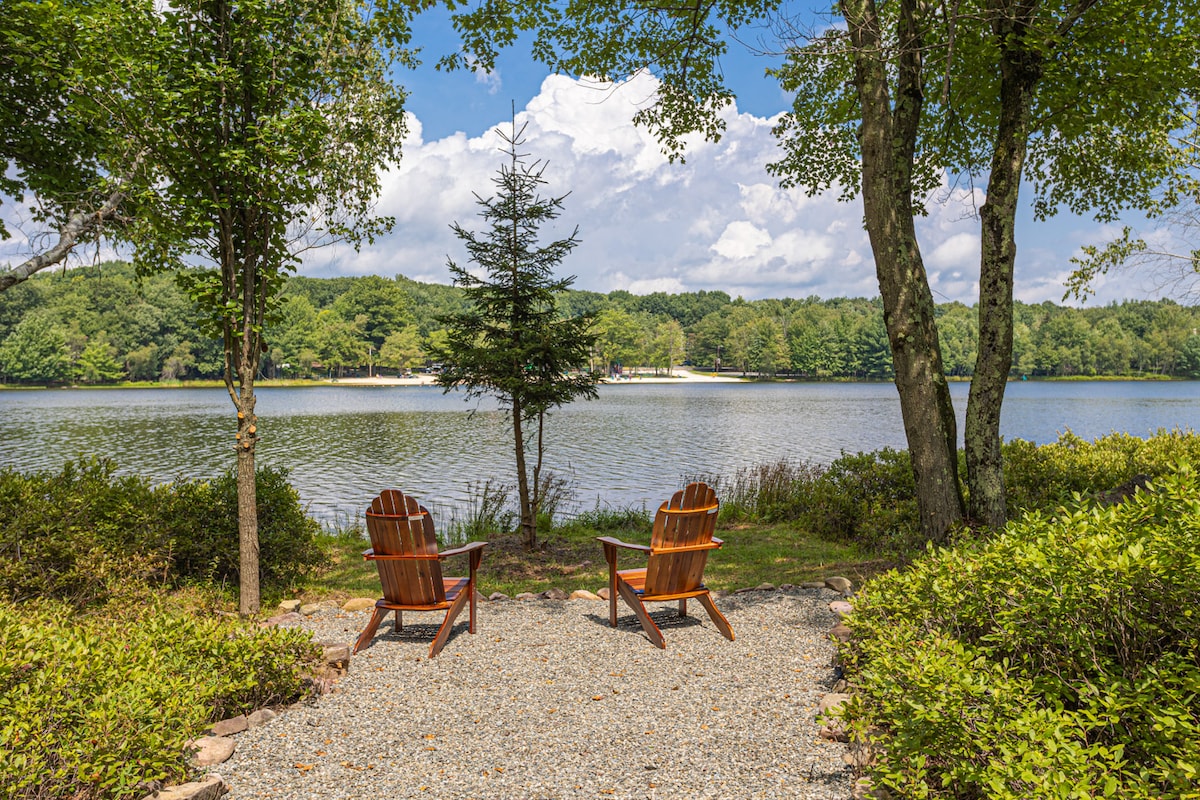 Two wooden chairs sit on a gravel area overlooking a calm lake. Lush greenery surrounds the seating, framed by trees and shrubs. The inviting space is set against a backdrop of water and a clear sky, creating a peaceful lakeside retreat.