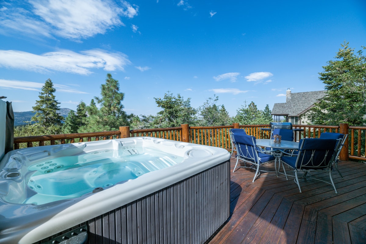 A hot tub is positioned on a spacious wooden deck, surrounded by trees and scenic mountain views. A dining table with four chairs is nearby, creating an inviting outdoor space. The clear sky fills the scene with natural light.