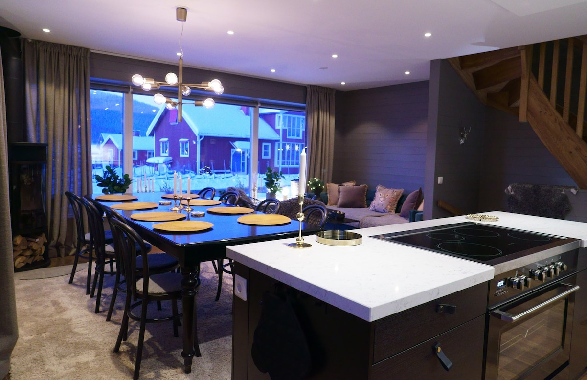 An open-plan dining area features a large table set for ten, surrounded by black chairs. A modern kitchen is equipped with sleek appliances. Panoramic windows provide a view of the snowy outdoors, while a cozy seating area with a sofa invites relaxation.